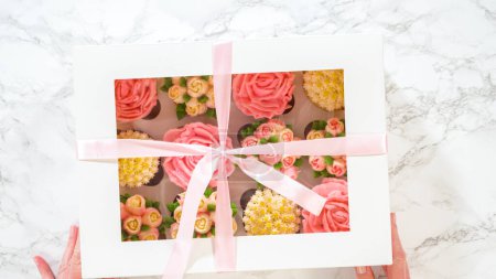 Photo for Flat lay. Held by a woman, a white paper box reveals gourmet cupcakes, beautifully decorated with buttercream frosting shaped into vibrant roses and tulips, a testament to culinary creativity. - Royalty Free Image