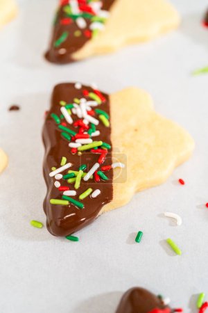Téléchargez les photos : Biscuits au beurre à moitié trempés dans du chocolat, saupoudrés de décorations festives rouges, vertes et blanches, posés sur du parchemin blanc à sertir. - en image libre de droit