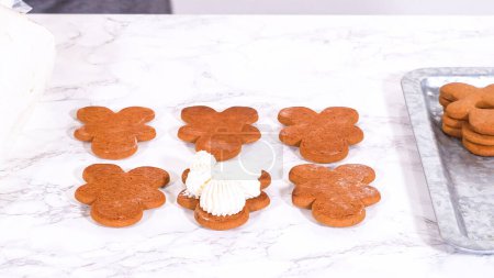 Gingerbread cookies await their second halves on a marble surface, each meticulously piped with buttercream to craft delightful sandwich treats. The precision of the piping adds a festive touch.