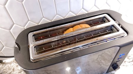 Photo for The image shows a sleek, contemporary toaster with a single slice of bread visible through its long slot, set against a tiled kitchen backsplash, with active blue indicator lights. - Royalty Free Image