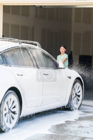 Foto de Denver, Colorado, Estados Unidos-1 de septiembre de 2023-Una joven ayuda con entusiasmo a lavar el automóvil eléctrico de las familias en su entrada suburbana. - Imagen libre de derechos