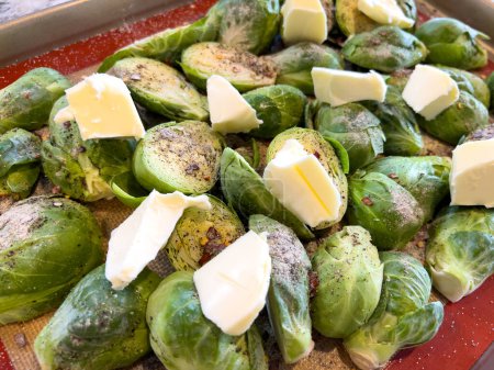Photo for A tray of whole Brussels sprouts is generously topped with butter pats and seasoned, set against a non-stick baking sheet, indicating preparation for a savory roast. - Royalty Free Image