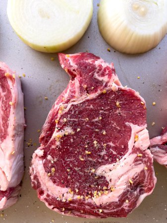 Three raw ribeye steaks with marbling, liberally seasoned with coarse spices, lie beside cut halves of onions on a kitchen tray, ready for cooking.