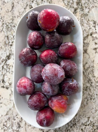 Photo for A plate of ripe, frosty plums sits prominently on a marble countertop in a sunlit kitchen with a crisp, white interior and neatly arranged appliances. - Royalty Free Image