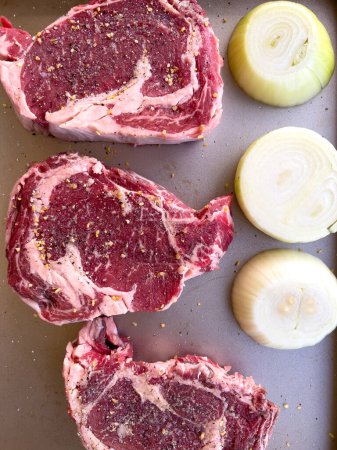Drei rohe Ribeye-Steaks mit Marmor, großzügig gewürzt mit groben Gewürzen, liegen neben geschnittenen Zwiebelhälften auf einem Küchentisch, fertig zum Kochen..