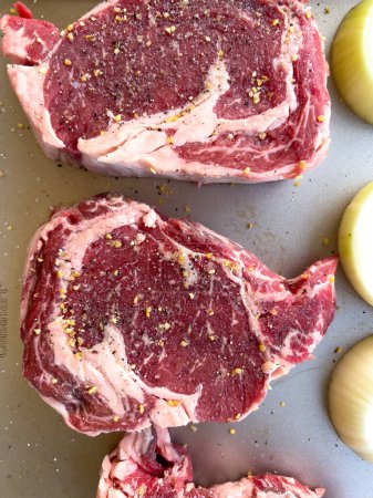 Trois steaks de côtelette crus au marbré, généreusement assaisonnés avec des épices grossières, se trouvent à côté de moitiés coupées d'oignons sur un plateau de cuisine, prêts à cuire.