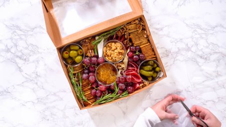 Photo for Flat lay. Hands are meticulously adding fresh red grapes to a bowl, complementing a beautifully arranged charcuterie box brimming with a variety of cheeses, olives, and cured meats, set against a - Royalty Free Image
