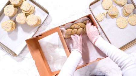 Téléchargez les photos : Pose plate. Avec précision, la femme arrange soigneusement les biscuits au sucre, remplis de saupoudres mélangées à la pâte, dans une boîte en papier brun rustique. - en image libre de droit