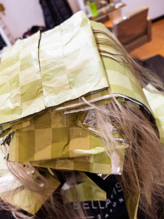 Strands of hair peek through a series of yellow foils meticulously arranged for a highlighting treatment. The salons ambiance is reflected in the background, where focused stylists perform their craft