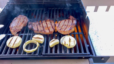 Ribeye steaks are sizzling alongside golden grilled onions on a barbecue grill, with wisps of smoke hinting at the flavorful feast being prepared. 