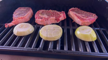 Des steaks de côtelette rôtissent aux côtés d'oignons grillés dorés sur un barbecue, avec des brins de fumée évoquant le festin savoureux en préparation.. 