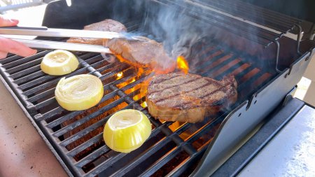 Ribeye steaks are sizzling alongside golden grilled onions on a barbecue grill, with wisps of smoke hinting at the flavorful feast being prepared. 
