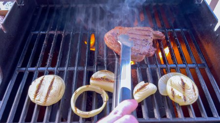 Ribeye-Steaks brutzeln neben goldenen gegrillten Zwiebeln auf einem Grill, und Rauchschwaden deuten auf das schmackhafte Festmahl hin, das zubereitet wird.. 