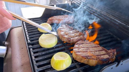 Des steaks de côtelette rôtissent aux côtés d'oignons grillés dorés sur un barbecue, avec des brins de fumée évoquant le festin savoureux en préparation.. 