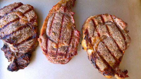 Three succulent ribeye steaks display perfect grill marks after being cooked, presented on a neutral surface, embodying the art of grilling.