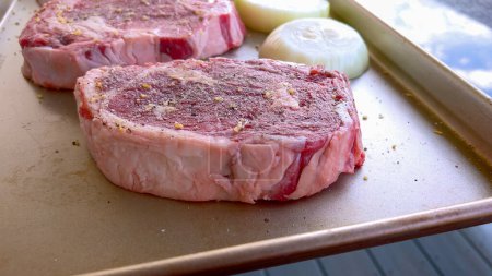 Dieses Bild zeigt drei rohe Ribeye-Steaks, großzügig gewürzt mit groben Gewürzen, neben Hälften frischer Zwiebeln auf einem Backblech, zubereitet für einen köstlichen Grillabend..