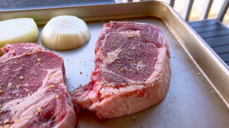 Téléchargez les photos : Cette image présente trois steaks crus, généreusement assaisonnés d'épices grossières, accompagnés de moitiés d'oignons frais sur une plaque à pâtisserie, préparés pour une délicieuse séance de grillades.. - en image libre de droit