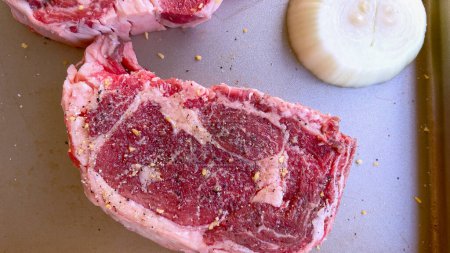Cette image présente trois steaks crus, généreusement assaisonnés d'épices grossières, accompagnés de moitiés d'oignons frais sur une plaque à pâtisserie, préparés pour une délicieuse séance de grillades..