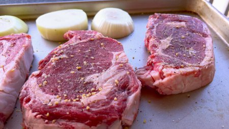 Photo for This image showcases three raw ribeye steaks, generously seasoned with coarse spices, alongside halves of fresh onions on a baking tray, prepared for a delicious grilling session. - Royalty Free Image