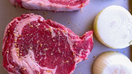 Téléchargez les photos : Cette image présente trois steaks crus, généreusement assaisonnés d'épices grossières, accompagnés de moitiés d'oignons frais sur une plaque à pâtisserie, préparés pour une délicieuse séance de grillades.. - en image libre de droit