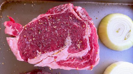 This image showcases three raw ribeye steaks, generously seasoned with coarse spices, alongside halves of fresh onions on a baking tray, prepared for a delicious grilling session.