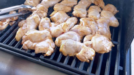 Une image en gros plan capturant le processus de grillage des morceaux de poulet marinés, avec une personne les retournant habilement pour assurer une cuisson uniforme sur un barbecue extérieur classique.
