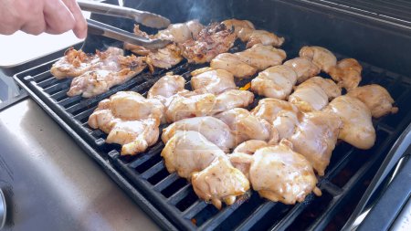 Ein Nahaufnahme-Bild, das den Prozess des Grillens marinierter Hühnerstücke einfängt, wobei eine Person diese fachmännisch umdreht, um sicherzustellen, dass sogar auf einem klassischen Grill im Freien gegrillt wird..