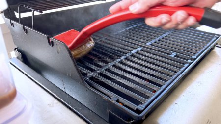 Une main utilise une brosse à griller rouge pour nettoyer les grilles noires d'un barbecue, s'assurant qu'il reste en parfait état pour la prochaine séance de grillage.