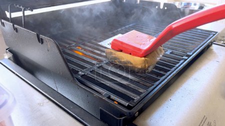 Téléchargez les photos : Une main utilise une brosse à griller rouge pour nettoyer les grilles noires d'un barbecue, s'assurant qu'il reste en parfait état pour la prochaine séance de grillage. - en image libre de droit
