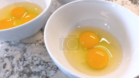 This image showcases three raw eggs cracked into a white bowl, set on a marble countertop, ready for mixing or cooking, highlighting the simplicity and beauty of basic cooking ingredients.