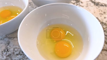 This image showcases three raw eggs cracked into a white bowl, set on a marble countertop, ready for mixing or cooking, highlighting the simplicity and beauty of basic cooking ingredients.