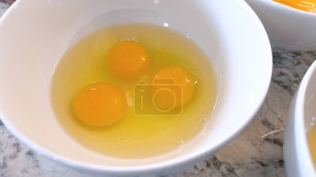 Dieses Bild zeigt drei rohe Eier, die in eine weiße Schüssel geknackt werden, die auf einer Marmorarbeitsplatte stehen und zum Mischen oder Kochen bereit sind, was die Einfachheit und Schönheit grundlegender Zutaten beim Kochen hervorhebt..