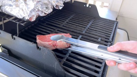 Esta imagen muestra el arte de asar a la parrilla, con tres filetes gruesos cocinando en una parrilla de barbacoa, con una fila de maíz envuelto en papel de aluminio en la mazorca de arriba, capturando una escena típica de una comida al aire libre abundante