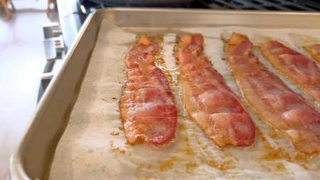 Téléchargez les photos : Cette image présente des bandes juteuses de cuisson au bacon dans le four sur une plaque de cuisson tapissée de parchemin, capturant la couleur brun doré attrayante et l'arôme attrayant comme il croustille. - en image libre de droit