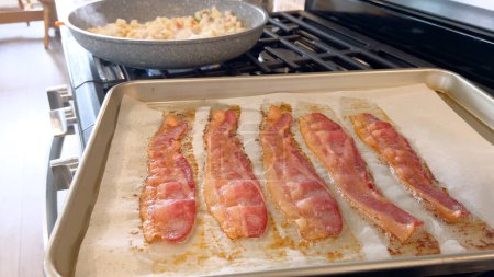 Cette image présente des bandes juteuses de cuisson au bacon dans le four sur une plaque de cuisson tapissée de parchemin, capturant la couleur brun doré attrayante et l'arôme attrayant comme il croustille.
