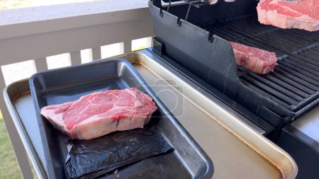 This image showcases the art of grilling, featuring three thick steaks cooking on a barbecue grill, with a row of foil-wrapped corn on the cob above, capturing a typical scene of a hearty outdoor meal