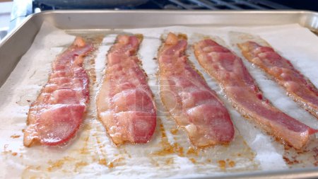 This image features juicy strips of bacon cooking in the oven on a parchment-lined baking tray, capturing the appealing golden-brown color and the enticing aroma as it crisps up.
