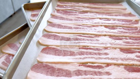 Bandes de bacon cru soigneusement disposées sur une plaque à pâtisserie, préparées pour la cuisson, capturant l'aspect frais et non cuit de cet ingrédient populaire avant qu'il ne devienne croustillant et doré.