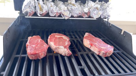Photo for This image showcases the art of grilling, featuring three thick steaks cooking on a barbecue grill, with a row of foil-wrapped corn on the cob above, capturing a typical scene of a hearty outdoor meal - Royalty Free Image