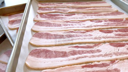 Bandes de bacon cru soigneusement disposées sur une plaque à pâtisserie, préparées pour la cuisson, capturant l'aspect frais et non cuit de cet ingrédient populaire avant qu'il ne devienne croustillant et doré.
