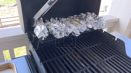 Photo for Several ears of corn wrapped in aluminum foil are lined up on a barbecue grill, slowly roasting to perfection, showcasing a popular and delicious method of cooking this classic side dish. - Royalty Free Image