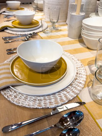 Ein schön arrangierter Esstisch mit modernem Geschirr mit einer auffallenden Farbpalette aus weißen und goldenen Tellern, ergänzt durch gestreifte Servietten und klare Gläser, die auf einem Holztisch für eine