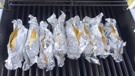 Photo for Several ears of corn wrapped in aluminum foil are lined up on a barbecue grill, slowly roasting to perfection, showcasing a popular and delicious method of cooking this classic side dish. - Royalty Free Image