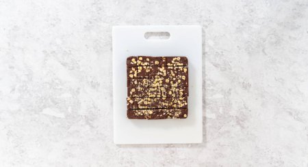 Flat lay. Cutting chocolate hazelnut fudge with a large kitchen knife into square pieces on a white cutting board.