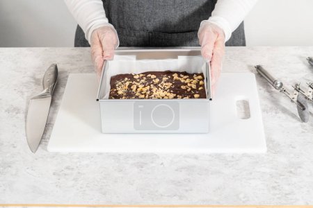 Removing chocolate hazelnut fudge from a square cheesecake pan lined with parchment.