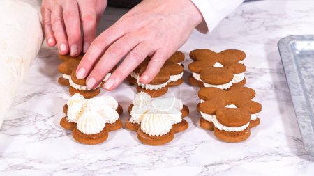 Las galletas de jengibre esperan sus segundas mitades en una superficie de mármol, cada una meticulosamente untada con crema de mantequilla para elaborar deliciosos bocadillos. La precisión de la tubería añade un toque festivo.