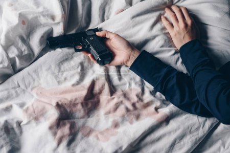 Photo for Cropped shot of woman lying on bed with gun in her hand and dry blood stains splash on bed sheet after her suicide herself. Suicide is the act of killing yourself caused by depression or illness. - Royalty Free Image