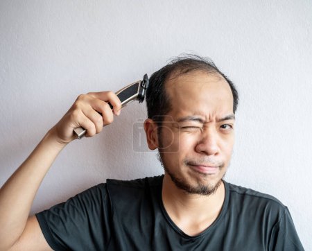 Portrait of Asian man trying to cut his hair by himself with electric hair clipper in covid-19 pandemic situation. People under quarantine are cutting their own hair from home.