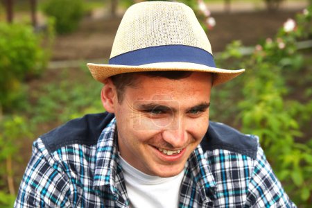 Foto de Un granjero sonriente con sombrero. Desenfoque agricultor en sombrero de trabajo en el campo con invernadero al aire libre. Retrato de joven caucásico guapo hombre feliz granjero en el campo y sonriendo. Fuera de foco. - Imagen libre de derechos