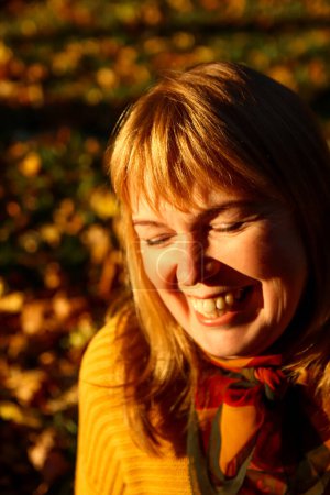 Foto de Retrato de cerca de una mujer alegre con una sonrisa radiante, disfrutando de las vibraciones de otoño en el parque, perfecto para revistas de estilo de vida, anuncios temáticos de otoño. Felicidad y bienestar. Fuera de foco. - Imagen libre de derechos